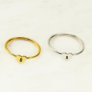 Puede incluir: Dos anillos, uno dorado y otro plateado, cada uno con un pequeño dije en forma de corazón. El anillo dorado tiene la letra "B" en el corazón, mientras que el anillo plateado tiene la letra "S".