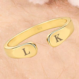 Op de afbeelding: Een goudkleurige open ring met een gladde, gepolijste afwerking, voorzien van de letter L aan het ene uiteinde en de letter K aan het andere uiteinde. De binnenkant van de ring is duidelijk gestempeld met het getal 925 voor sterling zilver.