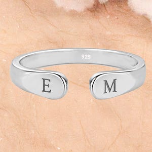 Puede incluir: Un anillo abierto de color plateado con acabado pulido y un sello 925 en la banda interior. Los dos extremos redondeados del anillo están grabados con las letras mayúsculas E y M, respectivamente, posicionadas hacia el exterior.