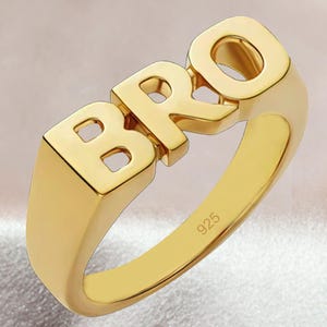 May include: A gold-toned ring featuring the letters BRO in a bold, block-style font across the top. The interior of the band is clearly stamped with the number 925, indicating the metal composition. The surface of the ring has a smooth, polished finish.