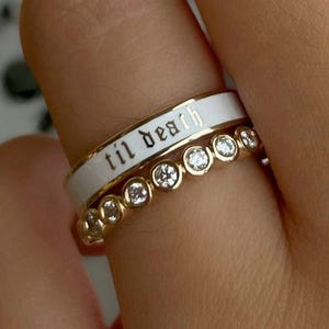 May include: Two gold rings. The top ring has a white band with the words "til death" in black script. The bottom ring is gold with clear stones. The rings are on a finger.