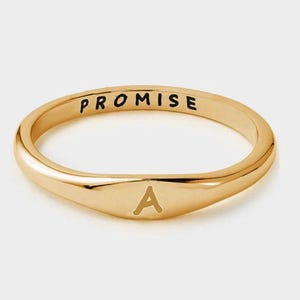 May include: A gold ring with the word "PROMISE" engraved on the top. The letter "A" is engraved on the front of the ring. The ring has a smooth, polished finish.