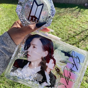 May include: A clear rectangular tray with a glittery finish. The tray features a photo of two men, one with red hair and the other with black hair. The tray also has silver flakes and a pink and purple gradient with the word "Proof" written in black. A separate silver glitter coaster with the BTS logo and the text "BTS" is also shown.