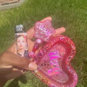 May include: A pink resin ashtray in a heart shape, a star-shaped keychain, and a lighter. The ashtray and keychain have the word "itzy" in pink. The lighter has a photo of a person.