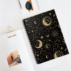Puede incluir: Una libreta espiral negra con un patrón celestial dorado. El patrón incluye lunas, estrellas y nubes.