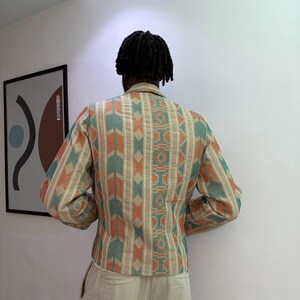 Puede incluir: Chaqueta beige con estampado geom&eacute;trico en verde azulado y naranja. La chaqueta tiene mangas largas y cuello. La persona lleva pantalones beige. La imagen est&aacute; tomada en interiores, con una obra de arte enmarcada en la pared.