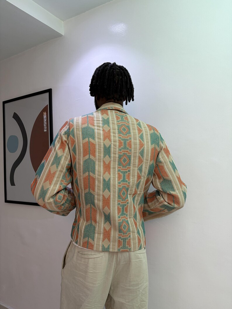 Puede incluir: Chaqueta beige con estampado geom&eacute;trico en verde azulado y naranja. La chaqueta tiene mangas largas y cuello. La persona lleva pantalones beige. La imagen est&aacute; tomada en interiores, con una obra de arte enmarcada en la pared.