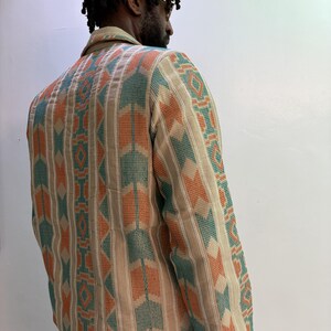 Puede incluir: Chaqueta beige con un patr&oacute;n geom&eacute;trico en verde azulado y naranja. La chaqueta tiene cuello y mangas largas. El patr&oacute;n es un dise&ntilde;o repetitivo de rayas verticales y formas de flecha. La chaqueta la lleva una persona.