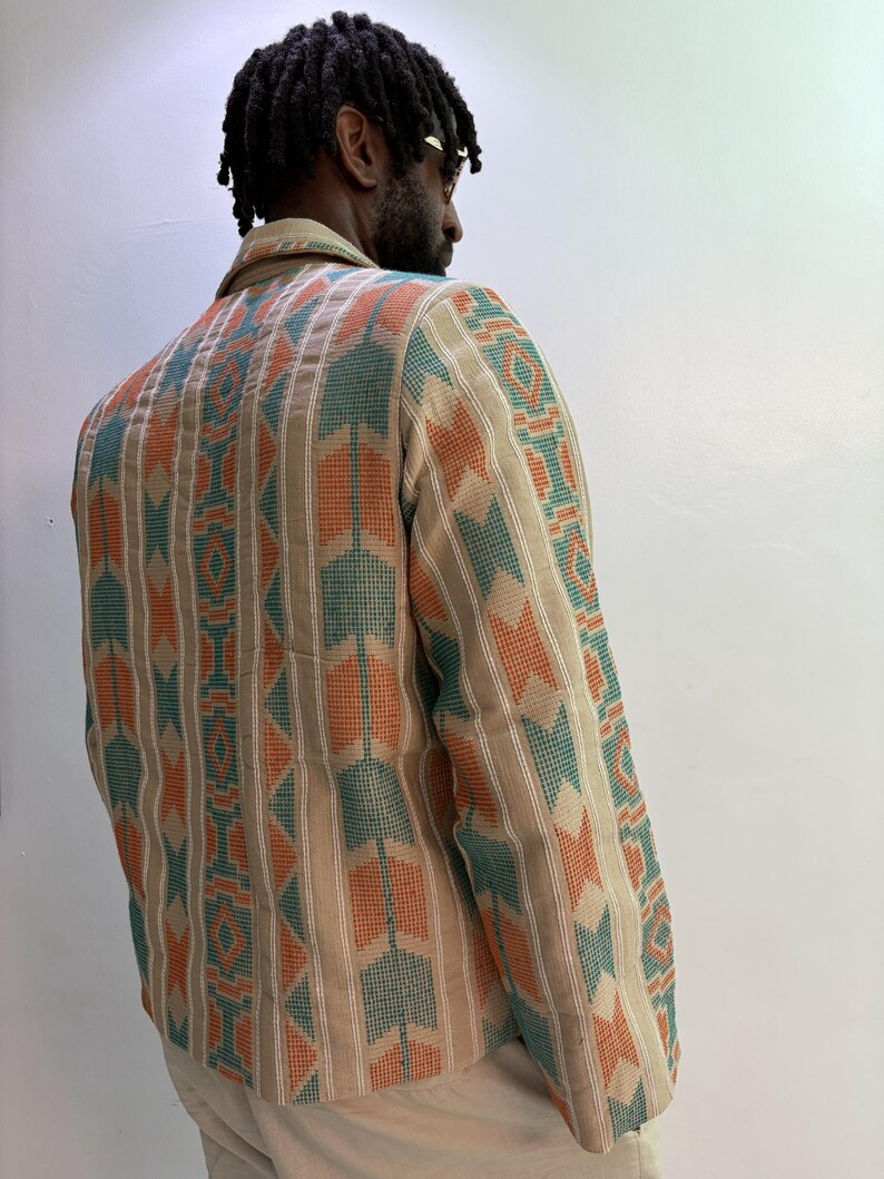 Puede incluir: Chaqueta beige con un patr&oacute;n geom&eacute;trico en verde azulado y naranja. La chaqueta tiene cuello y mangas largas. El patr&oacute;n es un dise&ntilde;o repetitivo de rayas verticales y formas de flecha. La chaqueta la lleva una persona.