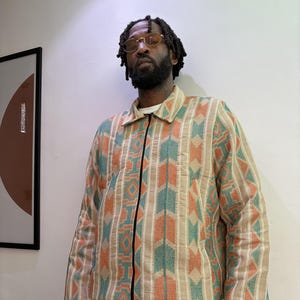 Puede incluir: Chaqueta beige con estampado geom&eacute;trico en naranja, turquesa y blanco. La chaqueta tiene una cremallera negra y cuello. La persona lleva una camisa blanca debajo y pantalones de color claro.