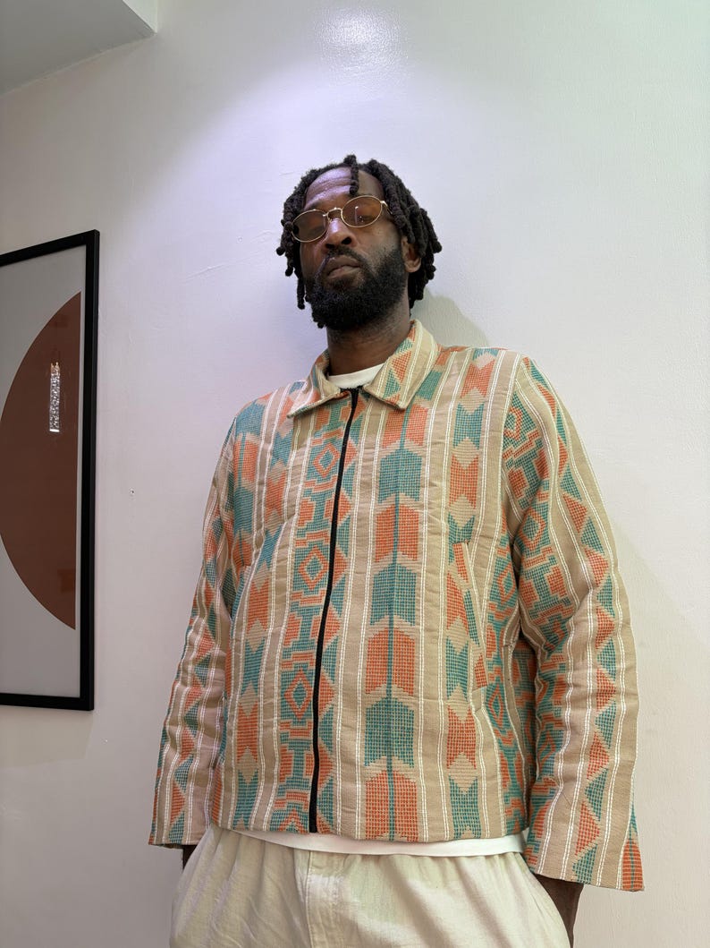 Puede incluir: Chaqueta beige con estampado geom&eacute;trico en naranja, turquesa y blanco. La chaqueta tiene una cremallera negra y cuello. La persona lleva una camisa blanca debajo y pantalones de color claro.