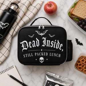 May include: Black lunch bag with the text "Dead Inside, Still Packed Lunch" in white gothic font, with bat and skull graphics. Includes a black water bottle, apple, sandwich, grapes, pretzels, and a notepad.