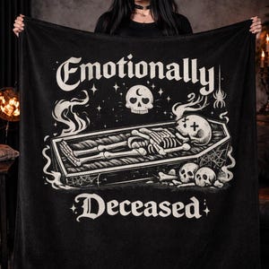 May include: Black blanket with white graphic text reading "Emotionally Deceased". The design features a skeleton in a coffin, skulls, and decorative elements. The blanket is held up, showcasing the design.