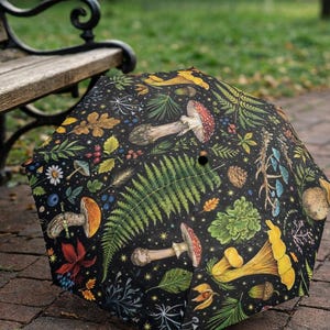 May include: A black umbrella with a colorful botanical pattern featuring mushrooms, ferns, berries, and leaves. The umbrella is open and resting on a brick surface, near a wooden bench.