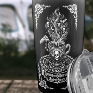 May include: Black travel mug featuring a white illustration of a skull-themed coffee cup, steam with skulls, and the text "Running on Caffeine and Ancient Curses." The mug has a clear lid and is on a wooden surface.
