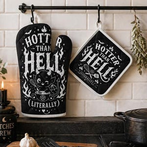 Könnte beinhalten: Schwarzer Ofenhandschuh und Topflappen-Set mit dem Text "HOTTER THAN HELL (LITERALLY)" und einer Teufelsgrafik. Ein schwarzes Schneidebrett mit dem Text "COOKING LIKE A BAD WITCH" hängt daneben. Küchenzubehör.