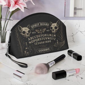 Puede incluir: Bolsa de cosméticos negra con un diseño de tablero de ouija, que incluye calaveras y el texto "SPIRIT BOARD". La bolsa está sobre una superficie de mármol con brochas de maquillaje y barras de labios.