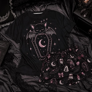 Può includere: Un set composto da una t-shirt nera a maniche corte e pantaloncini abbinati con una stampa grafica rosa. La maglietta mostra un pipistrello su una bara con una luna crescente e stelle. I pantaloncini presentano un motivo ripetuto di bare e pipistrelli.