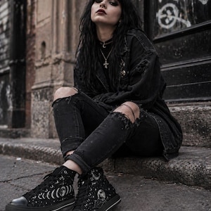 May include: Black high-top sneakers with a celestial moon and stars design. The sneakers have a black rubber sole and white stitching. The canvas shoes feature a lace-up closure and a round toe. The design includes phases of the moon and a full moon.