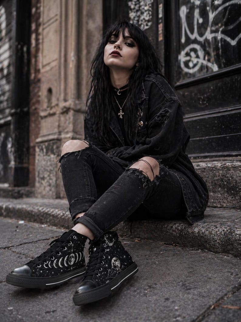 May include: Black high-top sneakers with a celestial moon and stars design. The sneakers have a black rubber sole and white stitching. The canvas shoes feature a lace-up closure and a round toe. The design includes phases of the moon and a full moon.