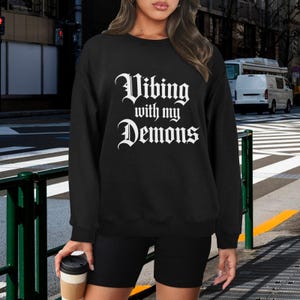 May include: Black crewneck sweatshirt with the text "Vibing with my Demons" in white gothic font. The sweatshirt is paired with black shorts. A person is holding a coffee cup.