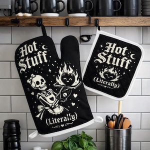 May include: Black oven mitt and pot holder set with the text "Hot Stuff (Literally)". The mitt features a skeleton server holding a tray with a flaming heart. The pot holder has a flaming heart design. Both have white trim.