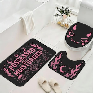 Può includere: Un set da bagno nero con dettagli rosa, tra cui un tappetino da bagno, un copriwater e un tappeto a forma di U. Il set presenta fiamme rosa, lune, stelle e la scritta "POSSESSED BUT MOISTURIZED". Il copriwater ha un design a forma di diavolo.