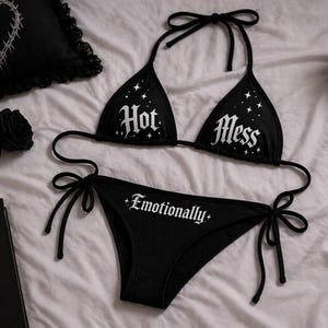 May include: Black bikini set with the words "Hot Mess" on the top and "Emotionally" on the bottom. The top has a triangle shape with tie straps. The bottom is a classic cut. The set is on a white bedspread.
