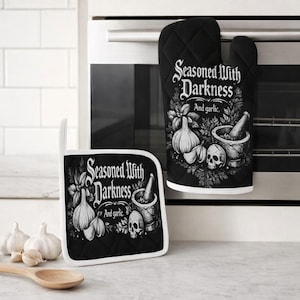 Könnte beinhalten: Schwarzes Ofenhandschuh- und Topflappen-Set mit gotischem Design. Der Text "Seasoned With Darkness And garlic" ist auf beiden Artikeln zu sehen. Das Design umfasst Abbildungen von Knoblauchzehen, einem Mörser und Stößel sowie einem Totenkopf.