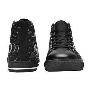 May include: Black high-top sneakers with a celestial design. The canvas uppers feature a night sky pattern with moons, stars, and bats. The sneakers have black laces, a black rubber sole, and a white stripe along the bottom.