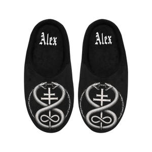 May include: Pair of black slippers with the name "Alex" in white gothic font. Each slipper features a silver emblem of two snakes around a cross and an infinity symbol.