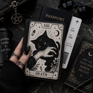 Op de afbeelding: Een zwarte paspoorthouder met een crèmekleurige tarotkaartillustratie van een zwarte kat die in een hand bijt, gelabeld met XIII en DEATH. Het ontwerp bevat hemelse motieven zoals sterren, een maansikkel en kristallen in een grafische stijl.