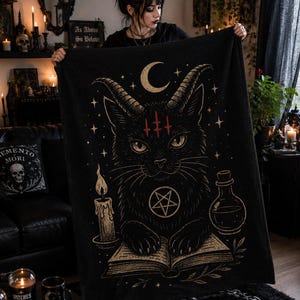 May include: A black rectangular throw blanket featuring a gold-toned illustration of a horned cat with a pentagram on its chest, resting its paws on an open book. The design includes a crescent moon, stars, a lit candle, and a potion bottle.