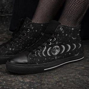 May include: Black high-top sneakers with a celestial design. The canvas shoes feature a black base with a pattern of stars, moons, and bats. The soles are black with a white stripe.