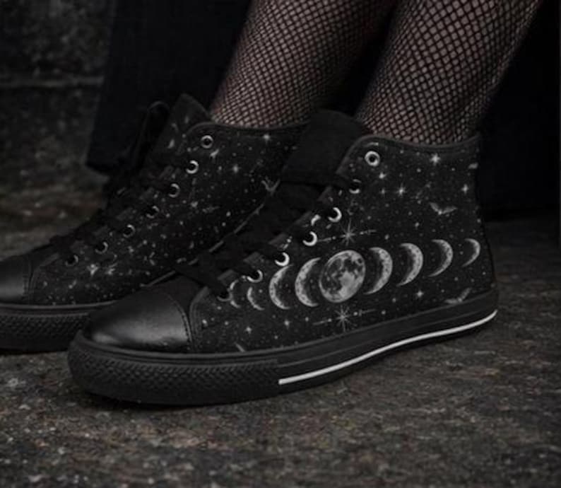 May include: Black high-top sneakers with a celestial design. The canvas shoes feature a black base with a pattern of stars, moons, and bats. The soles are black with a white stripe.