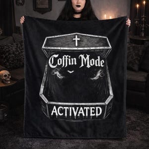 May include: Black blanket featuring a coffin design with the words "Coffin Mode Activated" in white. The blanket has a gothic theme, with a cross at the top of the coffin and a bat graphic.
