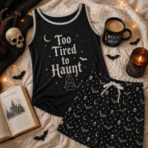 May include: Black pajama set with a tank top and shorts. The tank top has the text "Too Tired to Haunt" and a sleeping bat graphic. The shorts feature a pattern of bats, moons, and skulls. A mug with "Witch's Brew" is also visible.