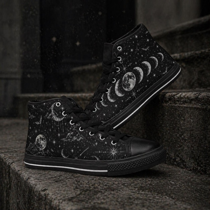 May include: Black high-top sneakers with a celestial design. The sneakers feature a black canvas upper adorned with a white moon phase and star pattern. The shoes have black laces, a black rubber toe cap, and a white stripe along the sole.