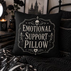 May include: A square black throw pillow featuring a white decorative frame with a skull at the top and a heart at the bottom. The text "EMOTIONAL SUPPORT PILLOW" is printed in the center in a distressed, serif font within the ornate border.