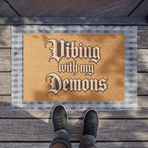 May include: A rectangular doormat with the phrase "Vibing with my Demons" in a gothic font. The mat has a tan center and a gray plaid border. Black shoes are visible in the foreground, suggesting the mat is at a doorway.