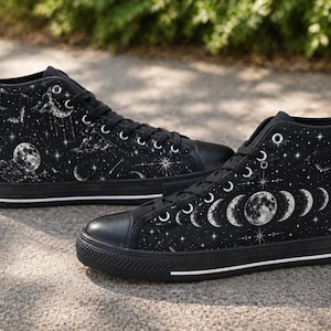 May include: Black high-top sneakers with a celestial design. The canvas shoes feature a black base with a white moon phases, stars, and clouds pattern. The sneakers have black laces, a black toe cap, and a white stripe on the sole.