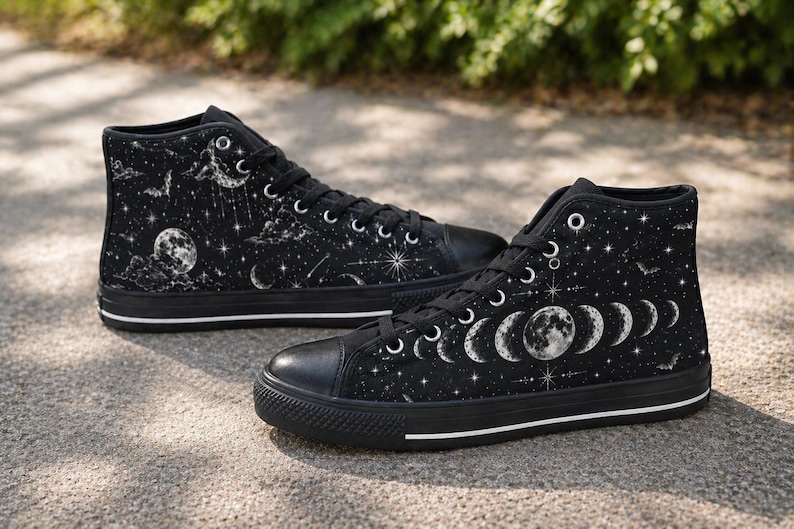 May include: Black high-top sneakers with a celestial design. The canvas shoes feature a black base with a white moon phases, stars, and clouds pattern. The sneakers have black laces, a black toe cap, and a white stripe on the sole.