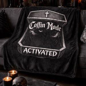 May include: Black plush blanket with a coffin graphic and the words "Coffin Mode Activated" in white. The blanket is draped over a gray sofa, with candles and a skull on a nearby table. Themed for Halloween.