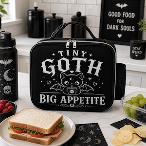 Könnte beinhalten: Schwarze Lunchtasche mit dem Text "TINY GOTH BIG APPETITE" und einer Illustration einer fledermausgeflügelten Katze. Eine schwarze Wasserflasche mit einem Fledermaus-, Mond- und Totenkopf-Design befindet sich neben der Tasche. Ein Sandwich, Obst und Snacks sind ebenfalls zu sehen.