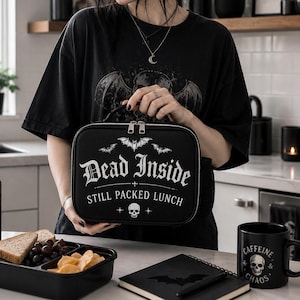 Pu&ograve; includere: Un portapranzo nero con la scritta "Dead Inside Still Packed Lunch" e grafiche di pipistrelli e teschi. L'immagine mostra anche una maglietta nera, una lattina di Liquid Death, una tazza nera con un teschio e un taccuino.