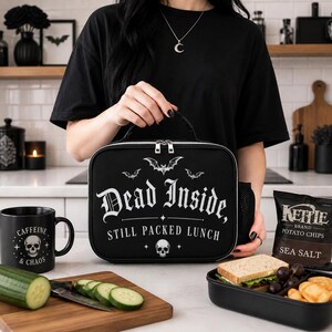 Pu&ograve; includere: Borsa per il pranzo nera con la scritta "Dead Inside, Still Packed Lunch" e grafiche di pipistrelli e teschi. &Egrave; visibile anche una tazza nera con "Caffeine & Chaos" e una grafica di teschio. Una busta di patatine e un panino sono anche nell'immagine.
