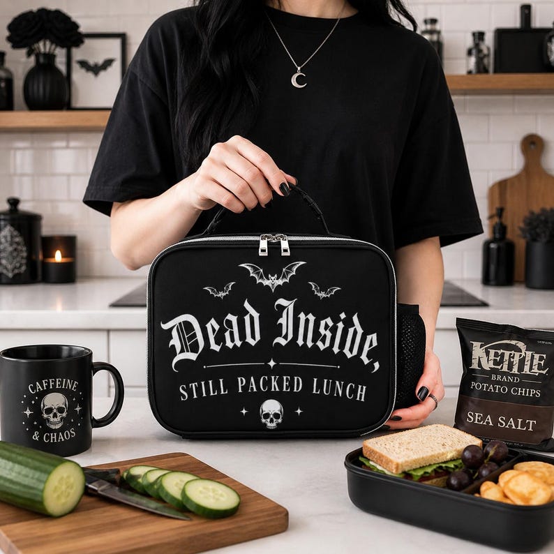 Pu&ograve; includere: Borsa per il pranzo nera con la scritta "Dead Inside, Still Packed Lunch" e grafiche di pipistrelli e teschi. &Egrave; visibile anche una tazza nera con "Caffeine & Chaos" e una grafica di teschio. Una busta di patatine e un panino sono anche nell'immagine.