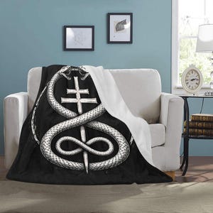 May include: Black and white throw blanket featuring a detailed illustration of two snakes intertwined around a cross and a dagger. The blanket is draped over a white sofa, with a white backing visible. The design has a gothic or occult theme.