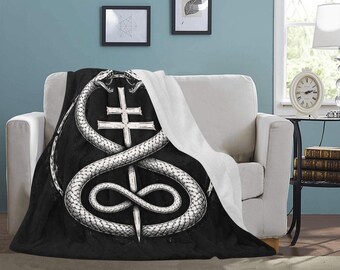 May include: Black and white throw blanket featuring a detailed illustration of two snakes intertwined around a cross and a dagger. The blanket is draped over a white sofa, with a white backing visible. The design has a gothic or occult theme.