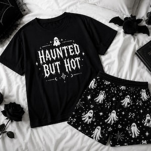 Può includere: Un set composto da una t-shirt a maniche corte e pantaloncini neri. La maglietta presenta una grafica bianca di un fantasma e la scritta "HAUNTED BUT HOT" con stelle. I pantaloncini hanno una stampa all-over di fantasmi bianchi, stelle, fiocchi e ragnatele.